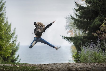 A woman floats in the air with her right arm and leg stretched and her left arm and leg bended. She wears a jacket, a pair of pants and a pair of sneakers, and carries a backpack. She is on a dirt path between tall tries and faces the ocean ahead.