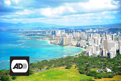 Panoramic view of Honolulu. The turquoise ocean bathes the beaches of the city. A large number of tall buildings rise along the coast. Superimposed over the image, in the lower left corner, the letters AD appear in black on a white background.