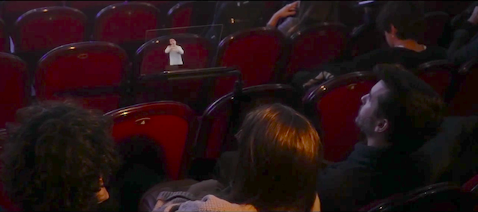 Several people sitting in a theater observe the transparent lectern in front of them showing the sign language interpreter.  The interpreter is wearing a light blue blouse.