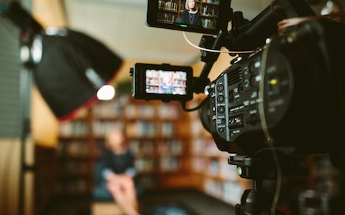 Indoors. A video camera focuses on a woman seated in front of a bookcase.