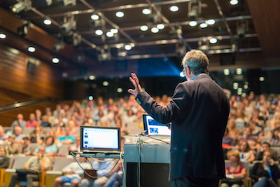 A speaker talks in front of an audience in a conference room.