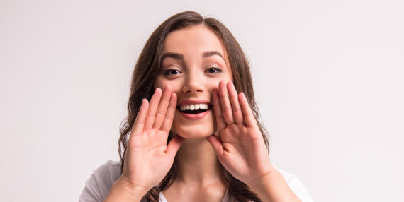 A woman looks forward, smiles and talks with her hands on the sides of her mouth.
