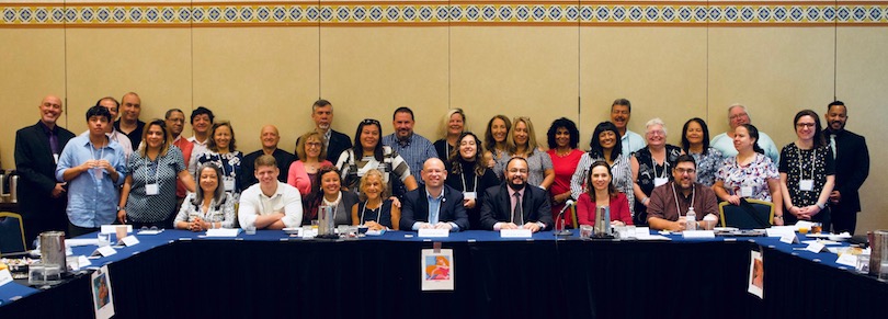 30 people smile behind a long table, half of them are seated and half of the are standing. 