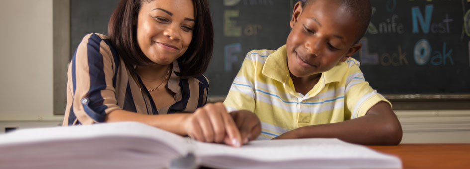 Classroom. A woman holds a boy's index finger over a page of a Braille book.
