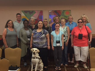Indoors, Judy and 11 people more stand and smile. A guide dog is seated in front.