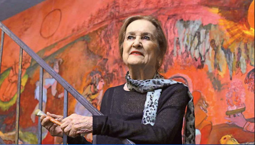 Rina Lazo, in her 80s, standing on a metal ladder in front of a mural. She holds a brush in her hand.