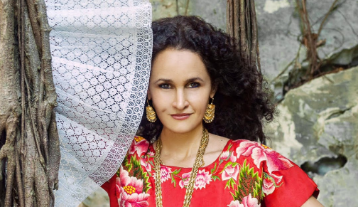 Susana Harp with long curly black hair poses next to a lace curtain that appears to be a fan.