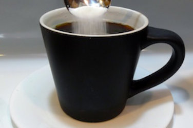 A cup of black coffee. White sugar falls in the coffee from a spoon.